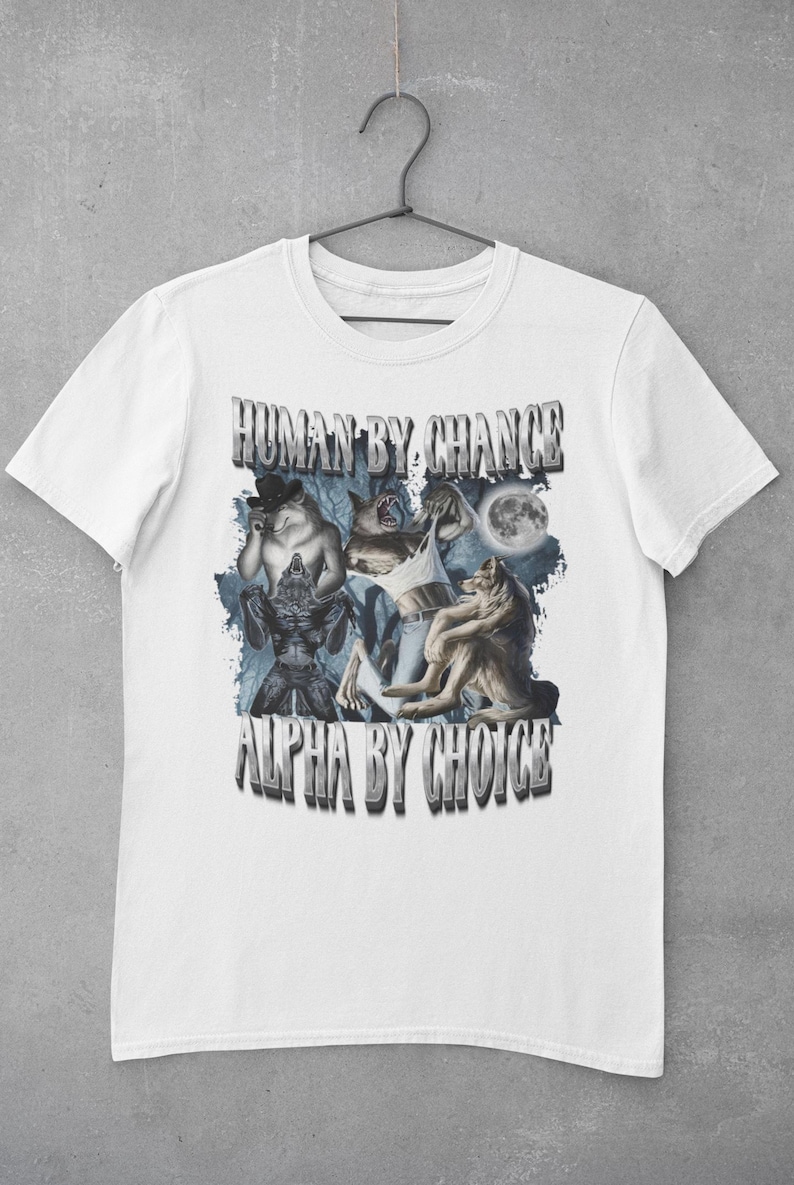 May include: A white short-sleeve t-shirt featuring a graphic print of multiple wolves and a full moon. The text "HUMAN BY CHANCE" appears at the top and "ALPHA BY CHOICE" at the bottom in a metallic, block-letter font. The design includes blue forest imagery.