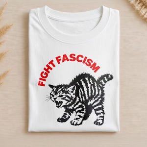 Puede incluir: Una camiseta blanca de cuello redondo con un gráfico negro de un gatito rayado bufando. Sobre el gatito, el texto "FIGHT FASCISM" está impreso en letras mayúsculas rojas y negritas. El diseño está centrado en la parte delantera de la prenda doblada.