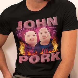 Op de afbeelding: Zwart T-shirt met een roze afbeelding van twee varkens die varkensmaskers dragen. De tekst "JOHN" staat boven de varkens en "PORK" eronder. De afbeelding is omringd door vlammen.
