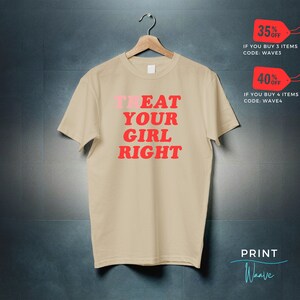 May include: Beige t-shirt with the text "TREAT YOUR GIRL RIGHT" in red and pink letters. The text is in a retro font style. The t-shirt is hanging on a wooden hanger against a gray background.
