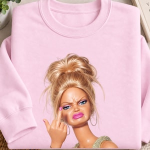 Op de afbeelding: Een lichtroze sweater met ronde hals en een grafische print van een blonde pop met een rommelige knot, die oorringen en een gouden top draagt. De pop heeft een fronsende gezichtsuitdrukking en steekt een middelvinger op.