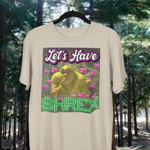 May include: A beige t-shirt with a green Shrek character image and the text "Let's Have Shrek" in pink and green neon lettering.