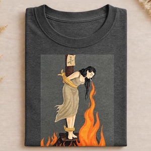 Op de afbeelding: Een donker antracietgrijs t-shirt met een grafische illustratie van een persoon met lang zwart haar die met gele touwen aan een houten paal is vastgebonden. De persoon rookt en oranje vlammen stijgen op. Er is een klein bordje bevestigd.