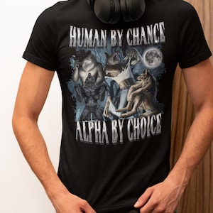 May include: A black short-sleeved t-shirt featuring a graphic print of three werewolf figures in a forest setting under a full moon. The text "HUMAN BY CHANCE" is printed above the graphic, and "ALPHA BY CHOICE" is printed below the graphic.