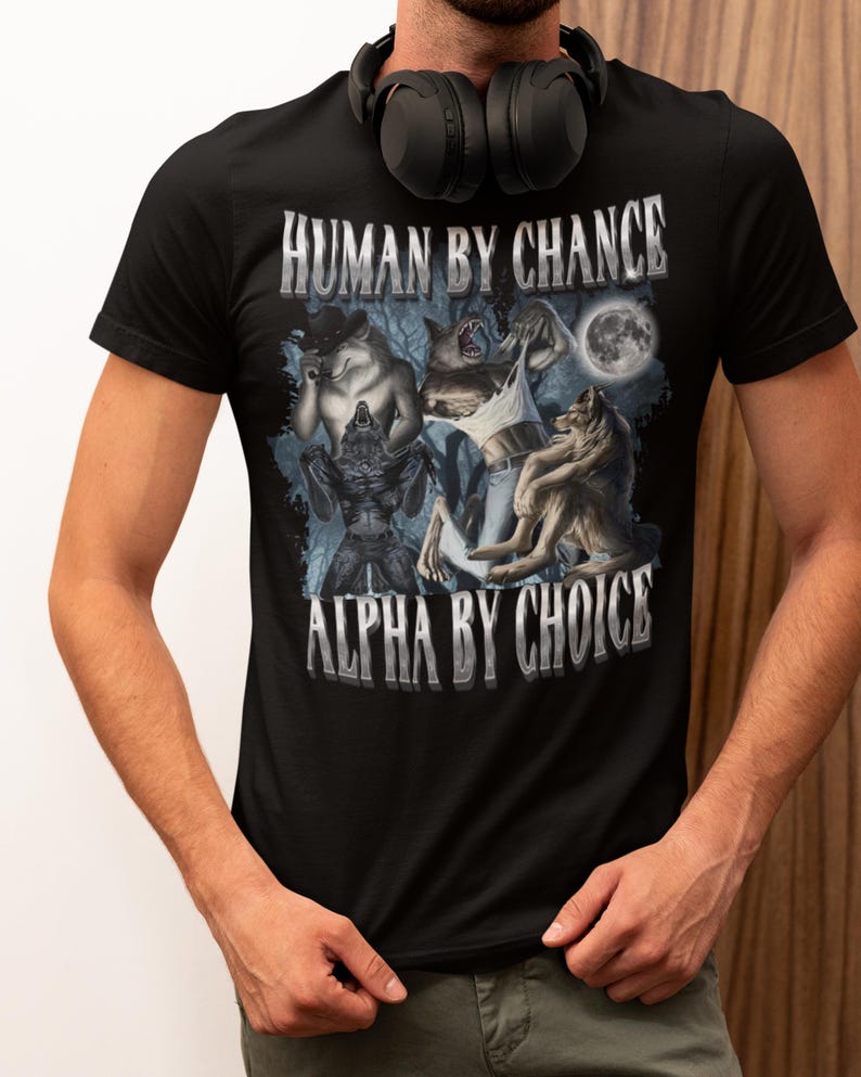 May include: A black short-sleeved t-shirt featuring a graphic print of three werewolf figures in a forest setting under a full moon. The text "HUMAN BY CHANCE" is printed above the graphic, and "ALPHA BY CHOICE" is printed below the graphic.