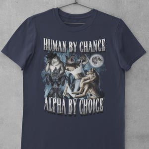 May include: A navy blue short-sleeve t-shirt featuring a graphic print of three werewolf figures in a forest setting under a full moon. The text "HUMAN BY CHANCE" is printed above the graphic, and "ALPHA BY CHOICE" is printed below the graphic.