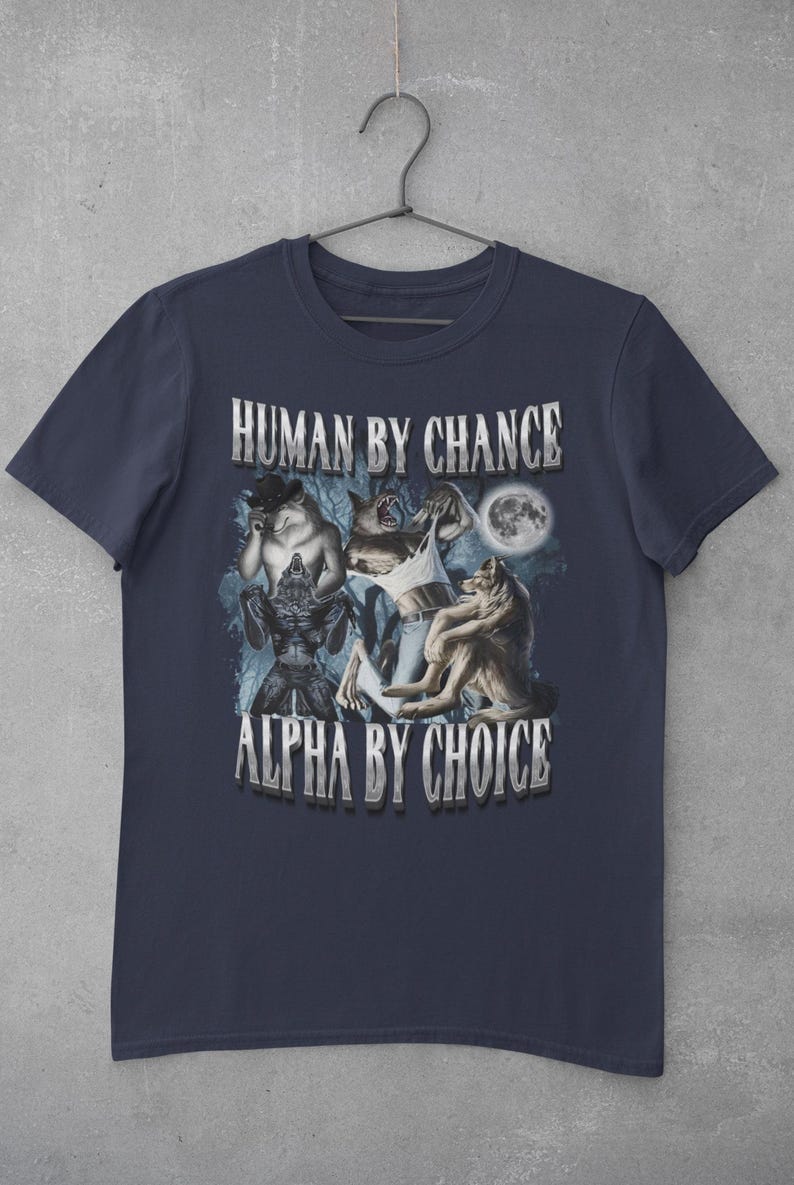 May include: A navy blue short-sleeve t-shirt featuring a graphic print of three werewolf figures in a forest setting under a full moon. The text "HUMAN BY CHANCE" is printed above the graphic, and "ALPHA BY CHOICE" is printed below the graphic.