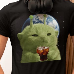 Op de afbeelding: Zwart t-shirt met een ontwerp van een groene alien-kat. De kat, met antennes, houdt een glas donkere drank met ijs vast, tegen een achtergrond van de aarde en de ruimte. De afbeelding heeft een grillige en speelse stijl.