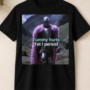 Könnte beinhalten: Ein schwarzes T-Shirt mit kurzen Ärmeln und einem Grafikdruck eines Ritters in silberner Rüstung, der einen langen violetten Pelzumhang trägt. Der Ritter steht in einer hügeligen Landschaft. Der Text "Tummy hurts Yet I persist" ist in Weiß über den Ritter gedruckt.