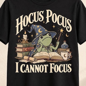 Könnte beinhalten: Ein schwarzes T-Shirt mit einer Grafik eines grünen Frosches, der einen blauen Zaubererhut mit Sternen trägt und einen Zauberstab hält. Der Frosch sitzt vor einem offenen Buch, Bücherstapeln, einer Kerze und einem Trank. Text HOCUS POCUS I CANNOT FOCUS.