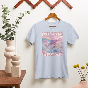 May include: Light blue t-shirt with a pink dolphin jumping over a rainbow in a cloud-filled sky. The text "LIVE, LAUGH LOBOTOMY" is printed on the shirt.