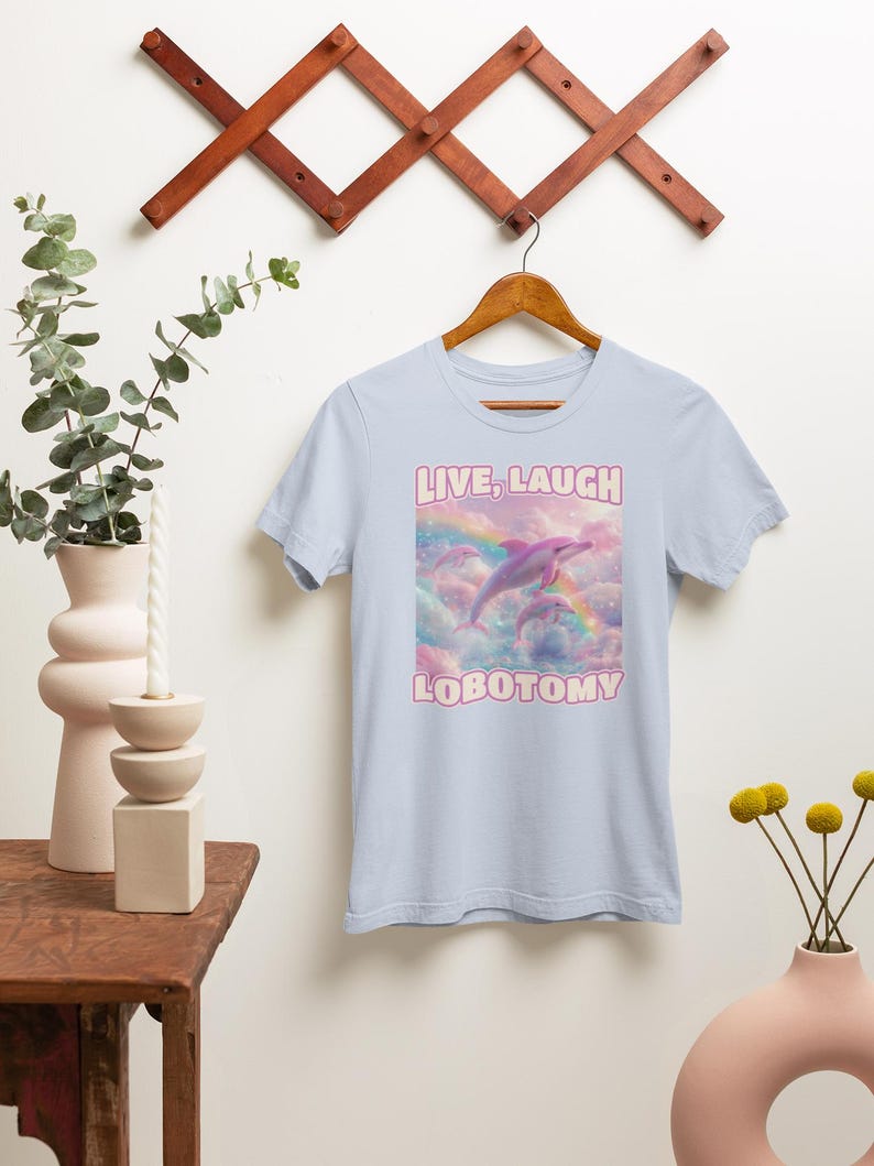 May include: Light blue t-shirt with a pink dolphin jumping over a rainbow in a cloud-filled sky. The text "LIVE, LAUGH LOBOTOMY" is printed on the shirt.