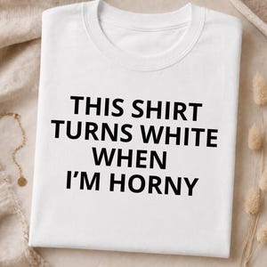Könnte beinhalten: Ein weißes kurzärmeliges T-Shirt mit Rundhalsausschnitt und schwarzem Text auf der Vorderseite, der lautet: THIS SHIRT TURNS WHITE WHEN I'M HORNY. Das Kleidungsstück ist ordentlich gefaltet und zeigt ein schlichtes Design mit fetter Schrift.