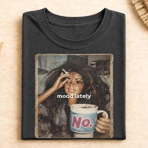 Könnte beinhalten: Ein schwarzes T-Shirt mit Rundhalsausschnitt und einem Grafikdruck einer Puppe mit zerzaustem dunklem Haar, die eine Zigarette hält. Der Text "mood lately" steht über einer Tasse, auf deren Vorderseite das Wort "No." in großen rosa Buchstaben steht.