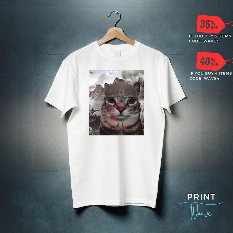 May include: White t-shirt with a graphic of a cat wearing a military helmet. The cat is looking directly at the viewer. The background is a blurry image of a war scene.