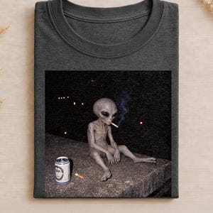 Op de afbeelding: Een donkergrijs T-shirt met ronde hals en een grafische print van een grijze alien die 's nachts op een richel zit. De alien rookt een sigaret met zichtbare blauwe rook en er staat een zilveren aluminium drankblikje naast op de richel.
