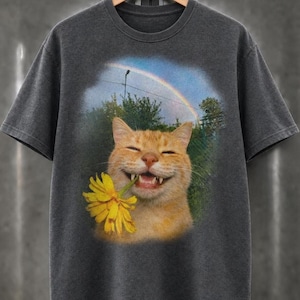 Könnte beinhalten: Ein anthrazitfarbenes Kurzarm-T-Shirt mit einem Grafikdruck einer orangefarbenen Katze, die die Augen geschlossen hat, lächelt und eine leuchtend gelbe Blume im Maul hält. Der Hintergrund zeigt einen Regenbogen am blauen Himmel über grünen Bäumen.