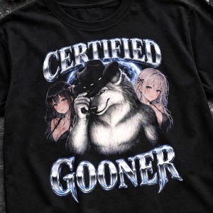May include: A black t-shirt featuring a graphic print of a wolf wearing a cowboy hat, flanked by two anime-style characters. The text "CERTIFIED" appears in metallic silver lettering above the wolf, and "GOONER" is printed below in the same style.