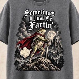 Puede incluir: Una camiseta gris carbón con el gráfico de una rana con capa roja que sostiene una espada sobre una roca. El texto "Sometimes I Just Be Fartin'" aparece impreso encima, con un castillo y una luna llena en el fondo de la ilustración.