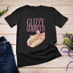 Puede incluir: Camiseta negra con el texto "Glizzy Gobber" en letras rosas y un dibujo animado de un perrito caliente con mostaza y un lazo rosa en la parte superior.