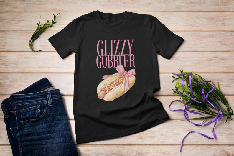 Puede incluir: Camiseta negra con el texto "Glizzy Gobber" en letras rosas y un dibujo animado de un perrito caliente con mostaza y un lazo rosa en la parte superior.