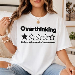 May include: A white short-sleeved t-shirt featuring the black text Overthinking above a graphic of five stars with only the first star filled in. Below the stars, the black text reads Endless spiral, wouldn't recommend.