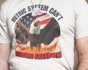 May include: White t-shirt featuring a graphic of an eagle with outstretched wings, soaring above an American flag with flames. The text "METRIC SYSTEM CAN'T MEASURE FREEDOM" is printed around the graphic.