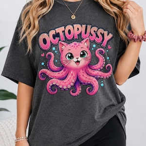 Può includere: Una t-shirt grigio antracite scuro con una stampa grafica di un gatto rosa con tentacoli di polpo. Il testo OCTOPUSSY è stampato in grandi lettere rosa sopra la creatura, accompagnato da piccole bolle blu e cuori rosa sul davanti.
