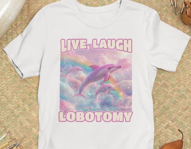 May include: White t-shirt with a pink graphic of three dolphins jumping through a rainbow in a cloudy sky. The text "LIVE, LAUGH LOBOTOMY" is printed in pink on the shirt.