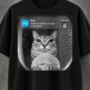 May include: A black t-shirt featuring a graphic print of a Ring doorbell notification that reads, "Ring There is motion at your Front Door 6m ago." The image shows a cat holding a fan of one hundred dollar bills in a fisheye lens perspective.