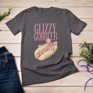 Puede incluir: Una camiseta gris oscuro con el texto "Glizzy Gobbler" impreso en rosa. Un hot dog de dibujos animados con mostaza y una cinta rosa est&aacute; impreso debajo del texto.