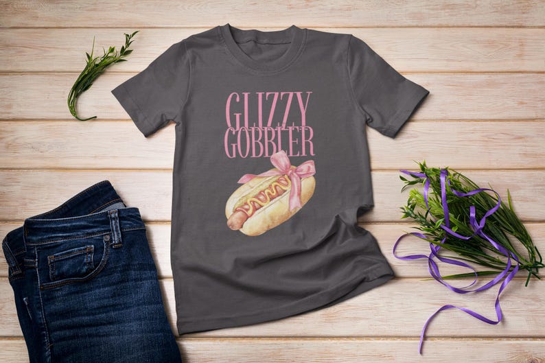 Puede incluir: Una camiseta gris oscuro con el texto "Glizzy Gobbler" impreso en rosa. Un hot dog de dibujos animados con mostaza y una cinta rosa est&aacute; impreso debajo del texto.