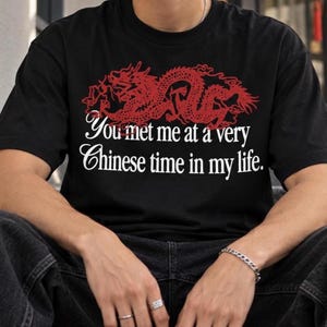 May include: A black short-sleeved t-shirt featuring a red graphic of a dragon positioned above white text that reads, You met me at a very Chinese time in my life. The shirt has a crew neckline and is made of a solid black fabric with a screen-printed design.