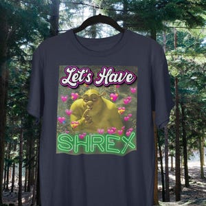 May include: A dark blue t-shirt with a green ogre character from the movie Shrek. The text "Let's Have Shrek" is printed on the shirt in a pink and white font.