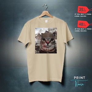May include: A beige t-shirt with a print of a cat wearing a military helmet. The cat is looking directly at the viewer. The background of the print is a blurry image of a war scene.