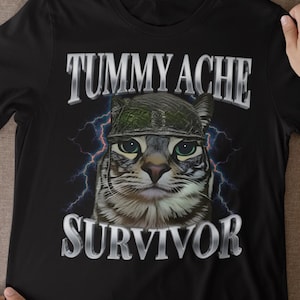 May include: Black t-shirt with a cat wearing a camouflage helmet. The words "TUMMY ACHE" and "SURVIVOR" are printed above and below the cat's image. Lightning bolts are on either side of the cat's head.