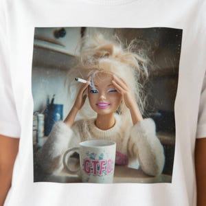 Può includere: T-shirt bianco con una grafica di una bambola Barbie con capelli biondi spettinati, che fuma una sigaretta e si tiene la testa. Davanti a lei c'è una tazza con le lettere "GTFO". L'immagine ha un effetto vintage leggermente sfocato.