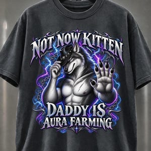 May include: A charcoal gray t-shirt featuring a graphic of an anthropomorphic wolf wearing a cowboy hat, with one hand raised and the other touching the hat brim. The text reads NOT NOW KITTEN DADDY IS AURA FARMING, surrounded by purple lightning.