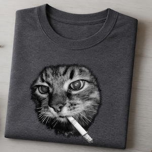 Peut inclure: Un t-shirt gris anthracite à col rond orné d'une impression graphique en noir et blanc représentant un chat tigré avec une cigarette à la bouche. Le visage du chat est centré sur la poitrine, montrant des détails de fourrure et des yeux.