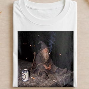 Könnte beinhalten: Ein weißes T-Shirt mit einem Grafikdruck eines bärtigen Zauberers mit spitzem Hut, der auf einem Felsvorsprung sitzt. Der Zauberer raucht eine Zigarette und hat einen Holzstab sowie eine silberne Getränkedose in seiner Nähe stehen.