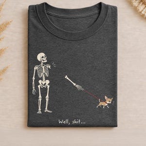 May include: A dark gray crew neck t-shirt featuring a white graphic of a skeleton missing an arm, with the detached arm being pulled away by a small brown chihuahua on a red leash. The text "Well, shit..." is printed in white below the graphic.