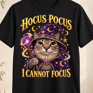 Può includere: Una t-shirt nera con la grafica di un gatto soriano che indossa un cappello e un mantello da mago viola. Il gatto tiene in mano un bastone luminoso. Il testo HOCUS POCUS è stampato sopra e I CANNOT FOCUS sotto in lettere dorate.