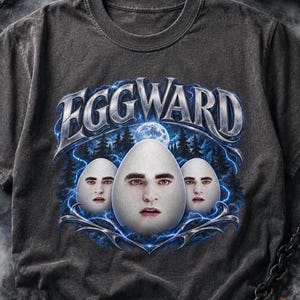 Könnte beinhalten: Ein anthrazitfarbenes T-Shirt mit einem Grafikdruck und dem Text EGGWARD in metallischer Schrift. Das Design zeigt drei weiße eiförmige Figuren mit menschlichen Gesichtern vor einem dunklen Waldhintergrund mit blauen Blitzen.