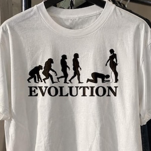 May include: A white short-sleeved t-shirt featuring a black graphic print of the human evolution sequence. The design shows a progression from primates to a human figure kneeling before a standing figure holding a leash, with the word EVOLUTION.
