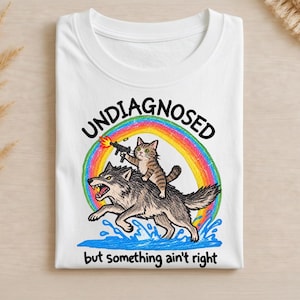 Może przedstawiać: Biały t-shirt z grafiką przedstawiającą pręgowanego kota jadącego na warczącym szarym wilku przez niebieską wodę. Kot trzyma strzelający pistolet maszynowy. Za nimi znajduje się kolorowa tęcza. Tekst głosi UNDIAGNOSED oraz but something ain't right.