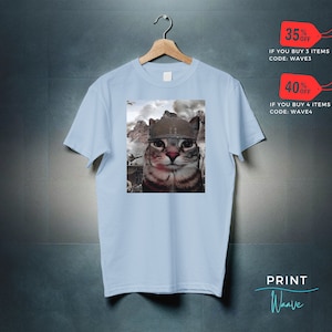 May include: A light blue t-shirt with a graphic of a cat wearing a military helmet. The cat is looking directly at the viewer. The background is a blurry image of a war scene.