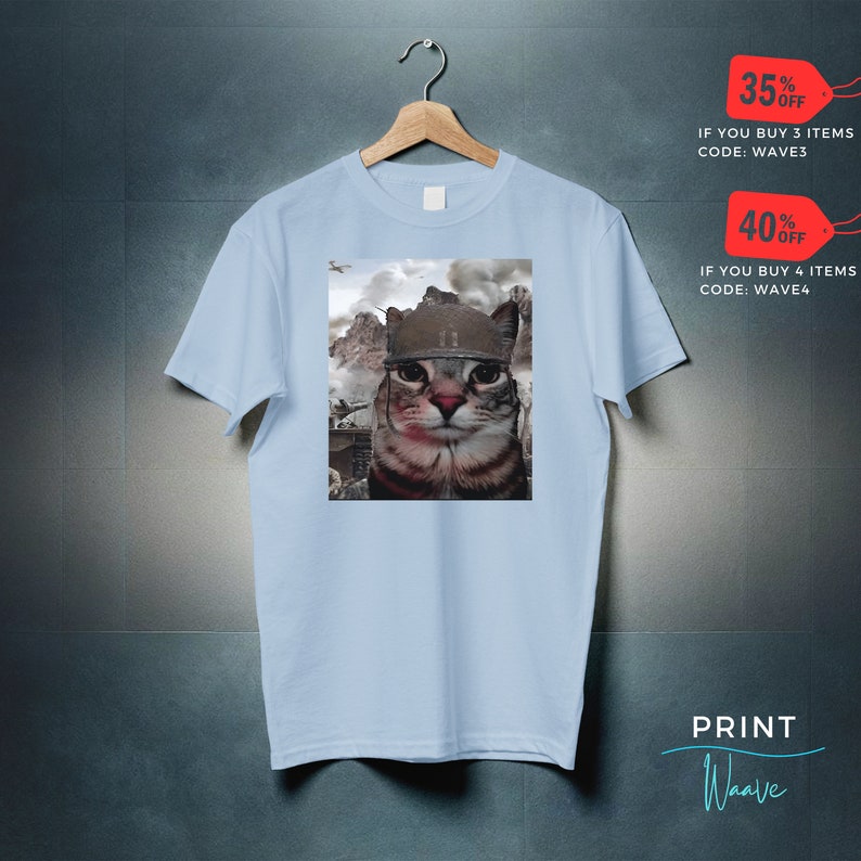 May include: A light blue t-shirt with a graphic of a cat wearing a military helmet. The cat is looking directly at the viewer. The background is a blurry image of a war scene.