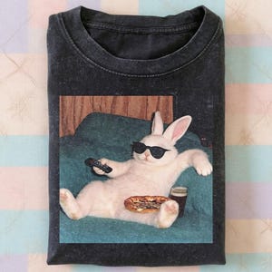 May include: Dark grey t-shirt with a graphic of a white rabbit wearing sunglasses, relaxing on a teal sofa, holding a remote, and with pizza and a drink. The rabbit is the main element.