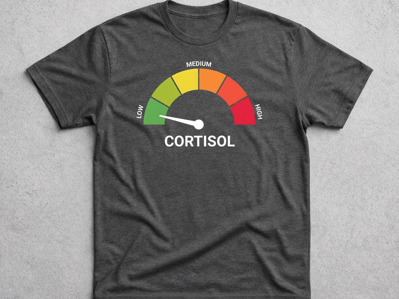 Pu&ograve; includere: T-shirt grigio scuro con grafica di un misuratore di cortisolo. Il misuratore ha una scala a colori da verde (basso) a rosso (alto), con un ago bianco. La parola "CORTISOL" &egrave; stampata in bianco sotto.