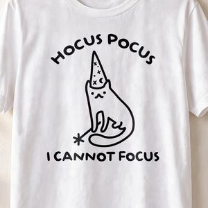 May include: A white short-sleeved t-shirt featuring a black line-art graphic of a cat wearing a wizard hat and holding a magic wand. The text HOCUS POCUS is printed above the cat, and the text I CANNOT FOCUS is printed below the cat.